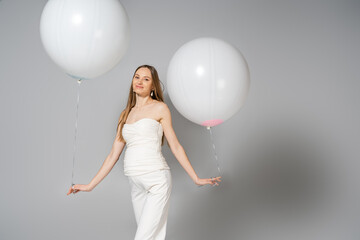 Positive and fashionable pregnant woman looking at camera while holding white festive balloons...