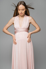 Fashionable and long haired pregnant woman in pink dress touching belly and looking down while standing isolated on grey, elegant and stylish pregnancy attire, sensuality, mother-to-be