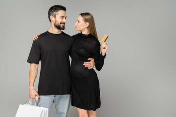 Smiling bearded man holding shopping bags and hugging stylish pregnant woman in black dress holding credit card isolated on grey, new beginnings and parenting concept, husband and wife