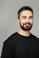 Portrait of positive bearded and brunette man in casual black t-shirt looking at camera while standing isolated on grey, masculine beauty concept, confident and charismatic