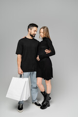 Man in jeans and t-shirt holding shopping bags and hugging trendy pregnant wife in black dress and standing together on grey background, new beginnings and shopping concept