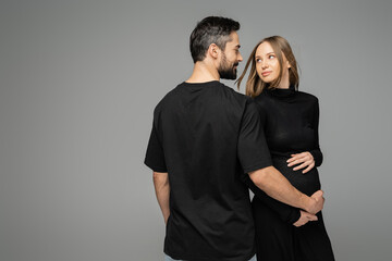 Smiling and bearded man in black t-shirt touching belly of pregnant stylish wife and looking at each other while standing isolated on grey, new beginnings and anticipation concept