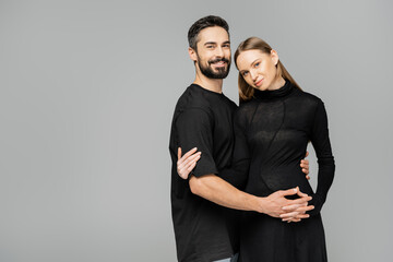 Positive and bearded husband in t-shirt looking at camera while hugging stylish pregnant wife in black dress isolated on grey, new beginnings and parenting concept
