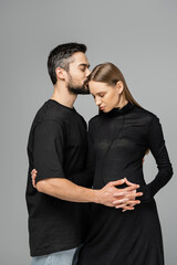 Bearded husband in t-shirt kissing and touching belly of stylish pregnant wife in black dress and standing isolated on grey, concept of expecting parents, happy family