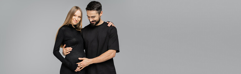 Cheerful bearded man in black t-shirt touching belly of fair haired and pregnant stylish wife in dress and hugging isolated on grey, growing new life concept, copy space, banner