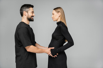 Side view of positive bearded man in t-shirt touching belly of pregnant wife in stylish black dress and looking at each other while standing isolated on grey, growing new life concept