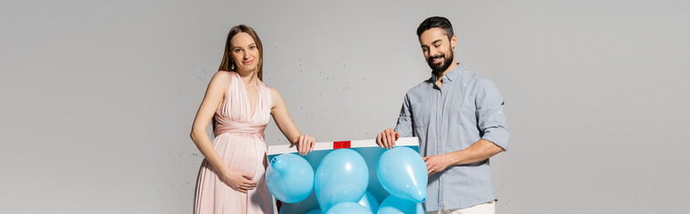 Elegant and pregnant woman opening gift box with blue balloons near cheerful husband and confetti during baby shower celebration on grey background, gender party, it`s a boy, banner