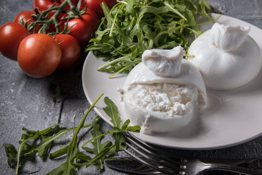 Cut Burrata cheese on a white plate with arugula and cherry tomatoes on a marble table, close up.