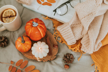 Burning candle, pumpkin decor and mug of lemon tea. Open book with eyeglasses and cozy sweater on bed. Autumn cozy home and hygge concept. Top view