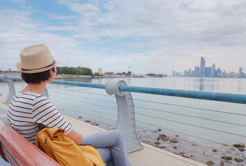 Obraz premium travel to the United Arab Emirates, Happy young asian female traveler with backpack and hat view to cityscape of downtown Abu Dhabi. Vacation and tourist destination concept.