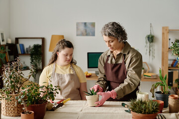 Woman in uniform teaching girl with down syndrome to plant plants at table in the room