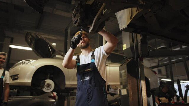Experienced mechanic staring at the car underside vehicle on a lift in service using a LED lamp. Two car mechanic diagnosing vehicle at the auto service. Workflow in a car workshop