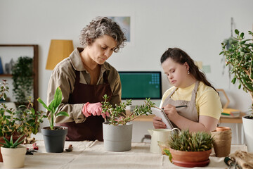 Florist transplanting green plants in pots while girl with down syndrome making notes in copybook