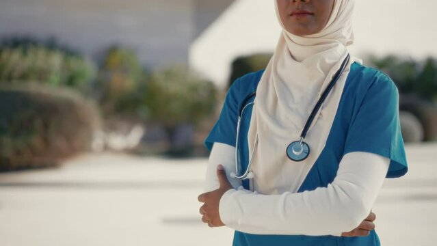 A young Muslim woman, a doctor in a hijab in uniform with a stethoscope, is standing on the street outside the hospital. Medicine and health care.
