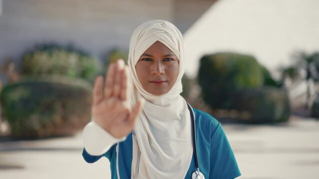 Young muslim woman in hijab doctor in uniform with stethoscope showing stop gesture with hand while standing on street near hospital building. Medicine and health care.