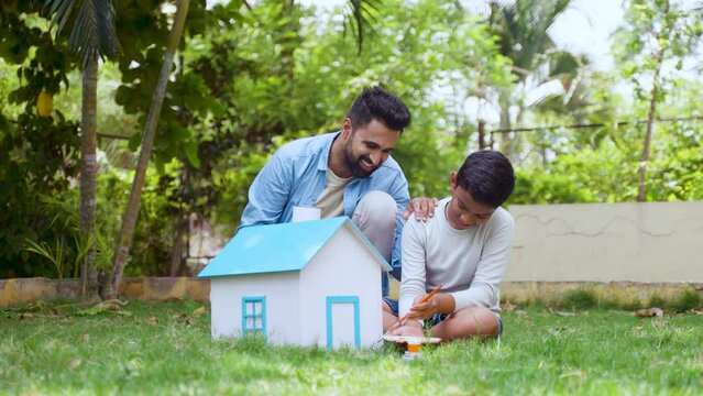 Happy indian father with kid painting cardboard toy house while sitting on park - concept of future planning, togetherness and fatherhood