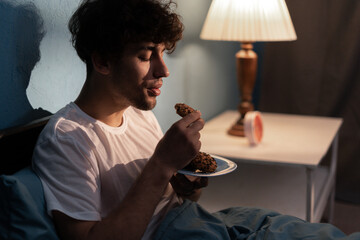 Stressed young man having a cookies at night. Unhappy tired depressed person eating during...