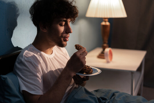 Stressed young man having a cookies at night. Unhappy tired depressed person eating during nighttime. Insomnia