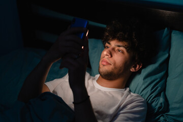 Insomnia, addiction concept. Sleepy exhausted man lying in bed using smartphone, can not sleep. Handsome guy in bed scrolling through social networks on mobile phone at night in dark - obrazy, fototapety, plakaty