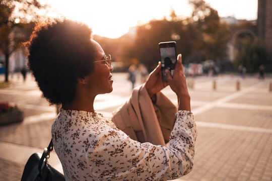 Smiling black woman, taking a photos of a beautiful city center, using a mobile phone.