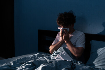 Handsome young man suffering cold and flu trying to sleep at night, guy sneezing into a tissue in bedroom. Illness colds or flu. Allergy or health issue concept. - obrazy, fototapety, plakaty