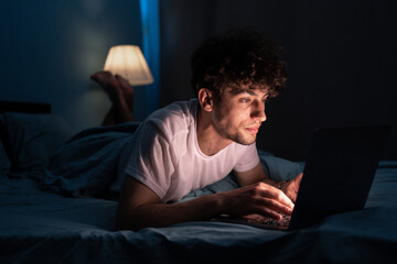 Young internet addict man networking concentrated late at night on bed with laptop in social media addiction or workaholic businessman - obrazy, fototapety, plakaty