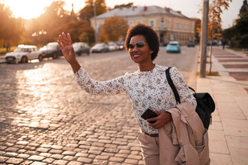 Smiling African tourist waving, calling a taxi ride, having a backpack, holding a coat.
