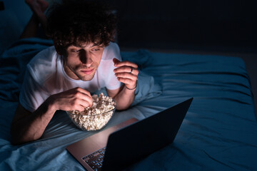 Young man eating popcorn and watching movie on laptop computer at night at home while lying on bed - obrazy, fototapety, plakaty