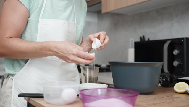 A woman is cooking in a bright kitchen. She cracks an egg and separates the egg white from the yolk into a glass.