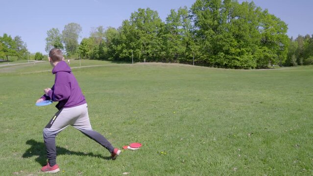Wide view of a teenage boy throwing a frisbee disc at full power in a green field with surrounding greenery and blue sky.