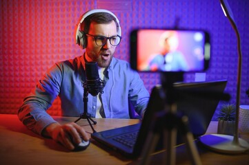 Naklejka na ściany i meble Young man recording or streaming podcast using microphone at his small broadcast studio. Content creator.