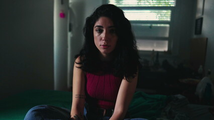 One young woman with serious expression portrait face closeup looking at camera inside a dark bedroom. Dramatic contrast of a female person in 30s