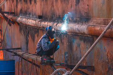 Welder on platform using arc welding machine to repairing the old rusty vessel hull at Shipyard area in evening time