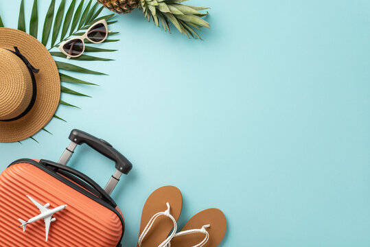 Summer adventure vibes. Top-down view of orange suitcase, miniature airliner, beach must-haves, glasses, sunhat, flip-flops, pineapple, palm leaves against pastel blue backdrop, space for text or ad