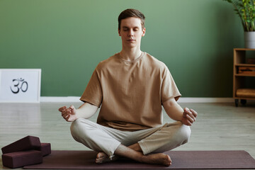 Fototapeta premium Front view portrait of young man meditating indoors sitting in lotus position with eyes closed, copy space