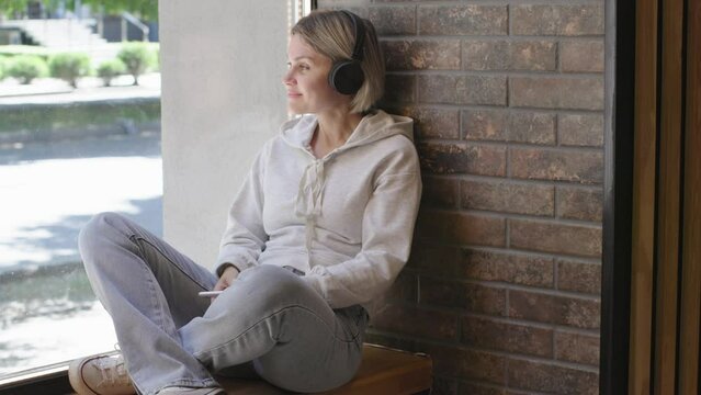 smiling female 30s 40s black headphones sitting windowsill city cafe using smartphone looking through panoramic window cars passing city summer day indoors. woman student tourist blue jeans lifestyle