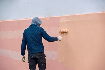 Man painting exterior building wall with paint roller brush outdoors. Worker manually painting pink wall, renovation facade works. Industrial painter at work, renovation of the building facade.