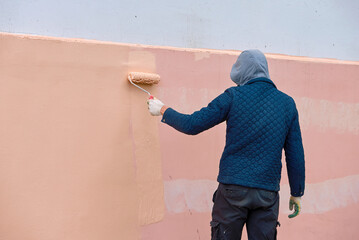 Man remove graffiti from building, painting wall with paint roller outdoors. Worker manually...