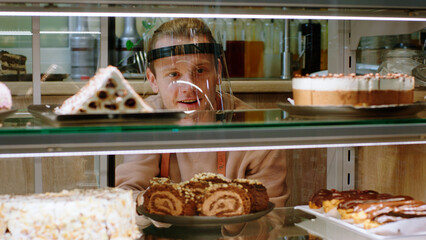 In front of the camera in the bakery cafe closeup smiling pretty salesman with protective mask in the pandemic of coronavirus arrange carefully the fresh and tasty deserts from the showcase fridge