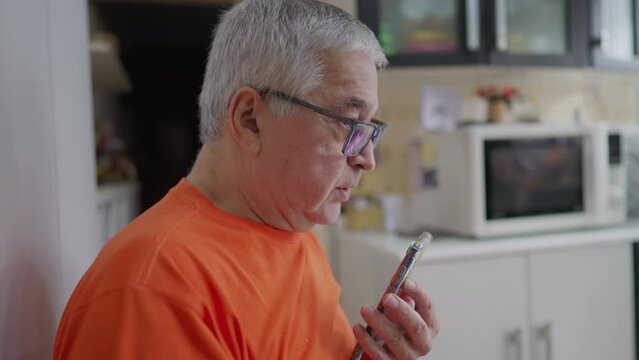 Senior man talking on phone sending voice message. Older male person in 70s in kitchen using modern technology