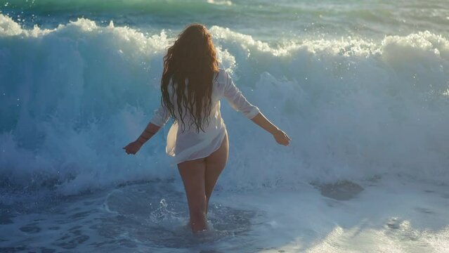 Young woman walking into waves of Mediterranean sea falling leaving in slow motion. Back view relaxed carefree female Caucasian tourist strolling in ocean at sunset