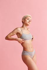 Portrait of young woman with short blonde hair, slim, fit body posing in grey cotton underwear against pink studio background