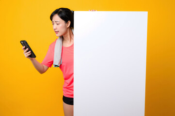 young asian sports fitness woman happy smile wearing pink sportswear standing behind the white blank banner or empty space advertisement board against yellow background. wellbeing application concept.