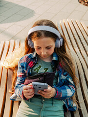 Blonde child in purple headphones checkered shirt listening to music podcast using smartphone bench outside city street urban lifestyle. Candid young girl learn online lesson enjoy audio book internet
