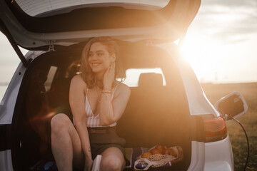 Young woman sitting in a car trunk, waiting for charging.