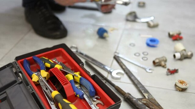 A male master plumber repairs a disassembled faucet from a bathroom, changes the cartridge and gasket. On the ceramic floor is a toolbox and a dismantled faucet.