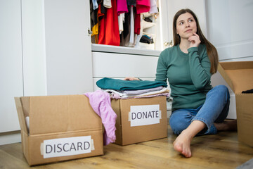 Woman sits on the floor next to cardboard boxes and thinks revision of her garment at home collects clothing for donation. Decluttering, orginizing clothes, personal stylist. Keep And Donate Fashion 