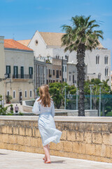 Fototapeta premium Blond hair girl walking on the ancient walls with Pontifical Basilica or Church of Saint Nicholas and palm trees on background in Bari old town, Puglia or Apulia, vertical