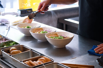 Bowls of ramen on a countertop in a kitchen with steam smoke of boiling noodles, hands put ingredients into a bowl with all supplies to preparing Japanese ramen to serve in a restaurant