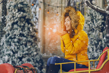 Pretty asian girl in yellow winter coat sitting on christmas rickshaw, hands making wishes, happy in front of pine tree grove with snow in winter.